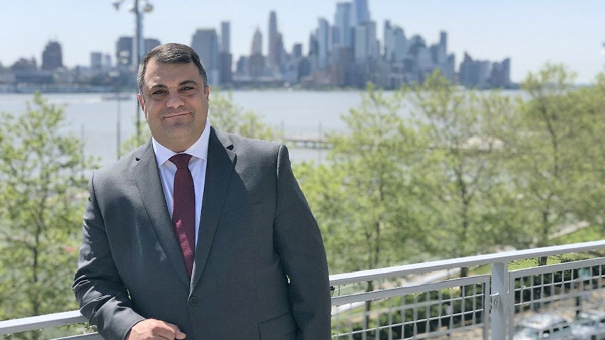 Chris Colla, in a gray suit and red tie, outside the Babbio Center with the New York City skyline behind him.