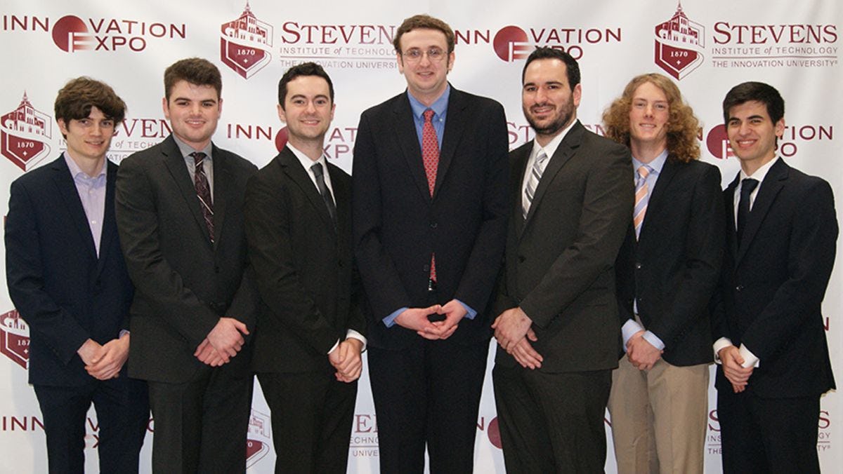 The seven students behind Coin Complex in front of the Stevens logo.