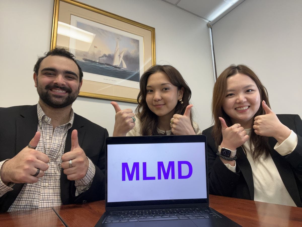 Three students each give two thumbs up on front of a laptop that displays "MLMD" on the screen. They are sitting at a wooden desk.