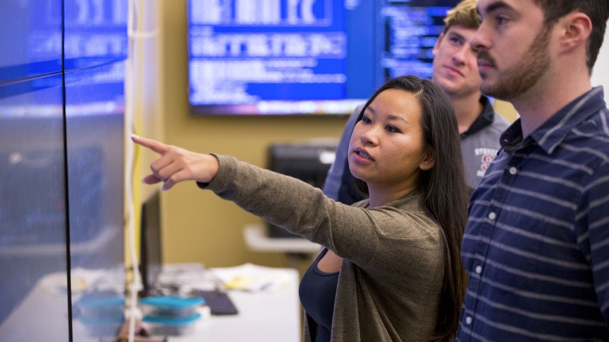 Student points to code on wall monitor while other students look on, impressed
