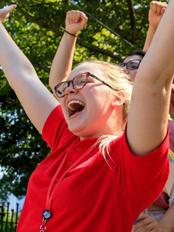 A female student in a red shirt throws her hands in the air, smiling.